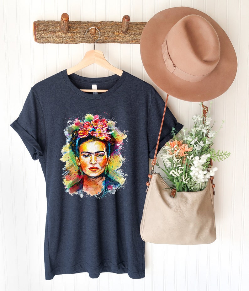 May include: A dark blue t-shirt with a colorful watercolor portrait of Frida Kahlo. The portrait features her signature flower crown and a colorful background.