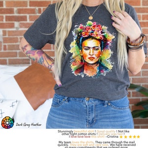 May include: Dark grey heather t-shirt featuring a colorful watercolor portrait of Frida Kahlo. The shirt has a crew neck and short sleeves. "Stunningly beautiful shirt! Great quality! Not like other tight cotton shirts! Delivery was fast! I love love love this shirt! - Cristina" and "My team loves the shirts. They came through the mail quickly. They are soft and very nice. We have received so many compliments that we ordered more. - Susan" are written below the image.