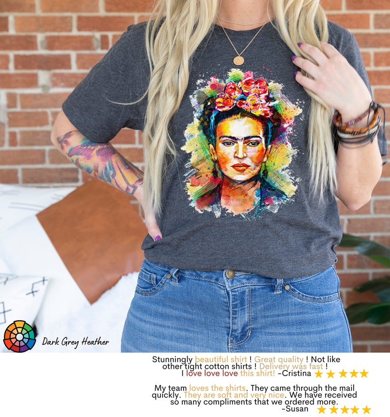 May include: Dark grey heather t-shirt featuring a colorful watercolor portrait of Frida Kahlo. The shirt has a crew neck and short sleeves. "Stunningly beautiful shirt! Great quality! Not like other tight cotton shirts! Delivery was fast! I love love love this shirt! - Cristina" and "My team loves the shirts. They came through the mail quickly. They are soft and very nice. We have received so many compliments that we ordered more. - Susan" are written below the image.