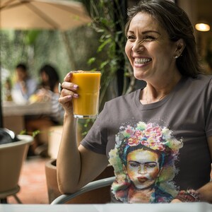 May include: A woman smiles while holding a glass of orange juice. She is wearing a grey t-shirt with a colorful painting of Frida Kahlo on it.