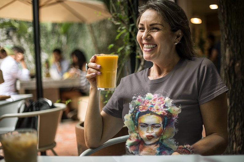 May include: A woman smiles while holding a glass of orange juice. She is wearing a grey t-shirt with a colorful painting of Frida Kahlo on it.