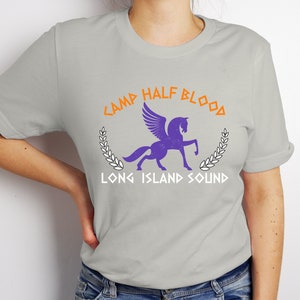 Puede incluir: Una camiseta gris con un caballo alado morado y el texto "Camp Half-Blood Long Island Sound" en naranja y blanco.