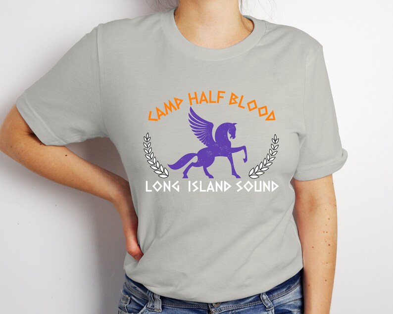 Puede incluir: Una camiseta gris con un caballo alado morado y el texto "Camp Half-Blood Long Island Sound" en naranja y blanco.