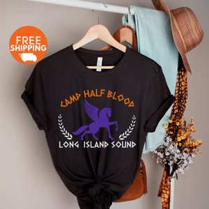 Puede incluir: Camiseta negra con un Pegaso morado y el texto "Camp Half Blood Long Island Sound" en naranja y blanco.