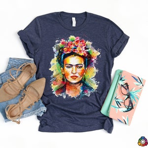 May include: A navy blue t-shirt with a colorful watercolor portrait of Frida Kahlo. The portrait features her signature flower crown and a splash of color.