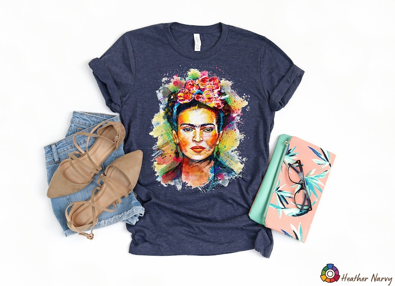 May include: A navy blue t-shirt with a colorful watercolor portrait of Frida Kahlo. The portrait features her signature flower crown and a splash of color.