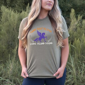 Puede incluir: Una camiseta verde con un pegaso morado y el texto "Camp Half-Blood Long Island Sound" en naranja y blanco.