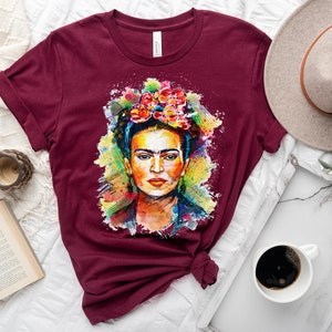 May include: A maroon t-shirt with a colorful watercolor painting of Frida Kahlo on the front. The painting features her signature flower crown and a colorful background.