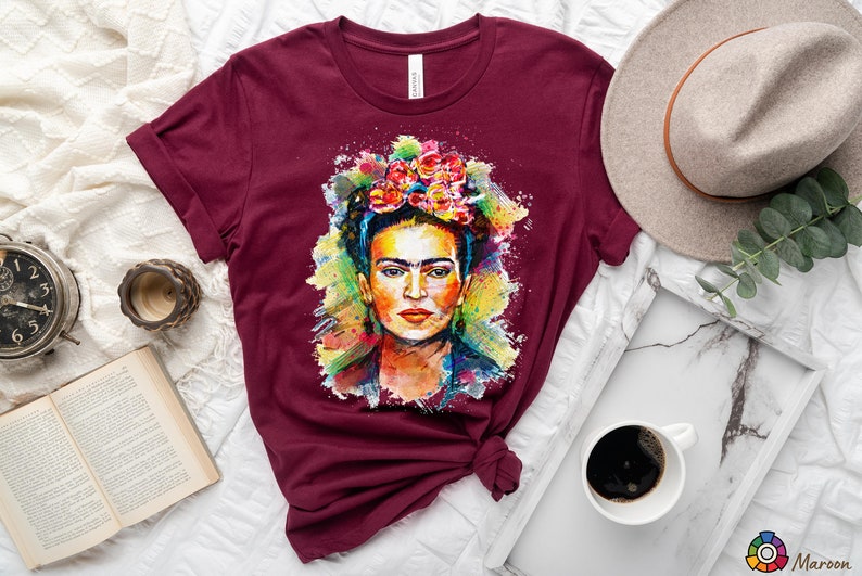 May include: A maroon t-shirt with a colorful watercolor painting of Frida Kahlo on the front. The painting features her signature flower crown and a colorful background.