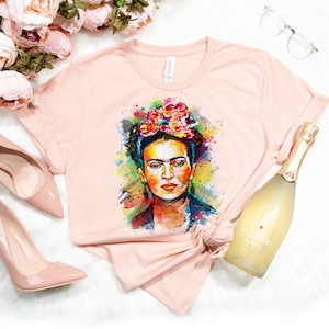 May include: A pink t-shirt with a colorful watercolor portrait of Frida Kahlo wearing a flower crown. The shirt is laid out on a white surface with a bottle of sparkling wine, a pair of glasses, and pink high heels.