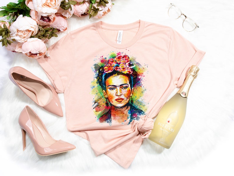May include: A pink t-shirt with a colorful watercolor portrait of Frida Kahlo wearing a flower crown. The shirt is laid out on a white surface with a bottle of sparkling wine, a pair of glasses, and pink high heels.