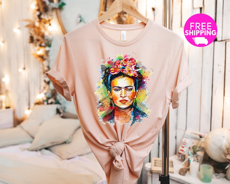 May include: A light pink t-shirt with a colorful watercolor illustration of Frida Kahlo. The illustration features her signature flower crown and a vibrant background.