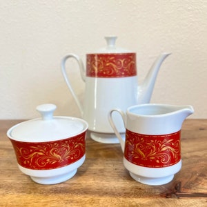 Puede incluir: Una tetera, una jarra de crema y un azucarero blancos con un diseño floral rojo y dorado. El juego está hecho de porcelana y tiene un aspecto vintage.