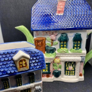 Puede incluir: Un juego de tetera y azucarero de cerámica azul y rosa con forma de casas con detalles florales. La tetera tiene un pico verde y una chimenea rosa. El azucarero tiene un techo azul y una chimenea blanca.
