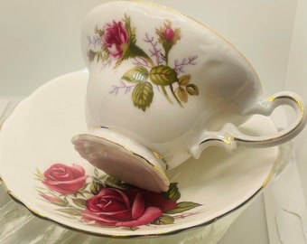 May include: A white teacup and saucer with a floral design featuring pink roses and green leaves. The cup and saucer have a gold trim.