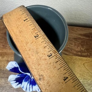 May include: A small, teal-colored ceramic vase with a blue and white flower accent. A wooden ruler is placed over the vase, measuring approximately 4 inches in height. The vase sits on a wooden surface.