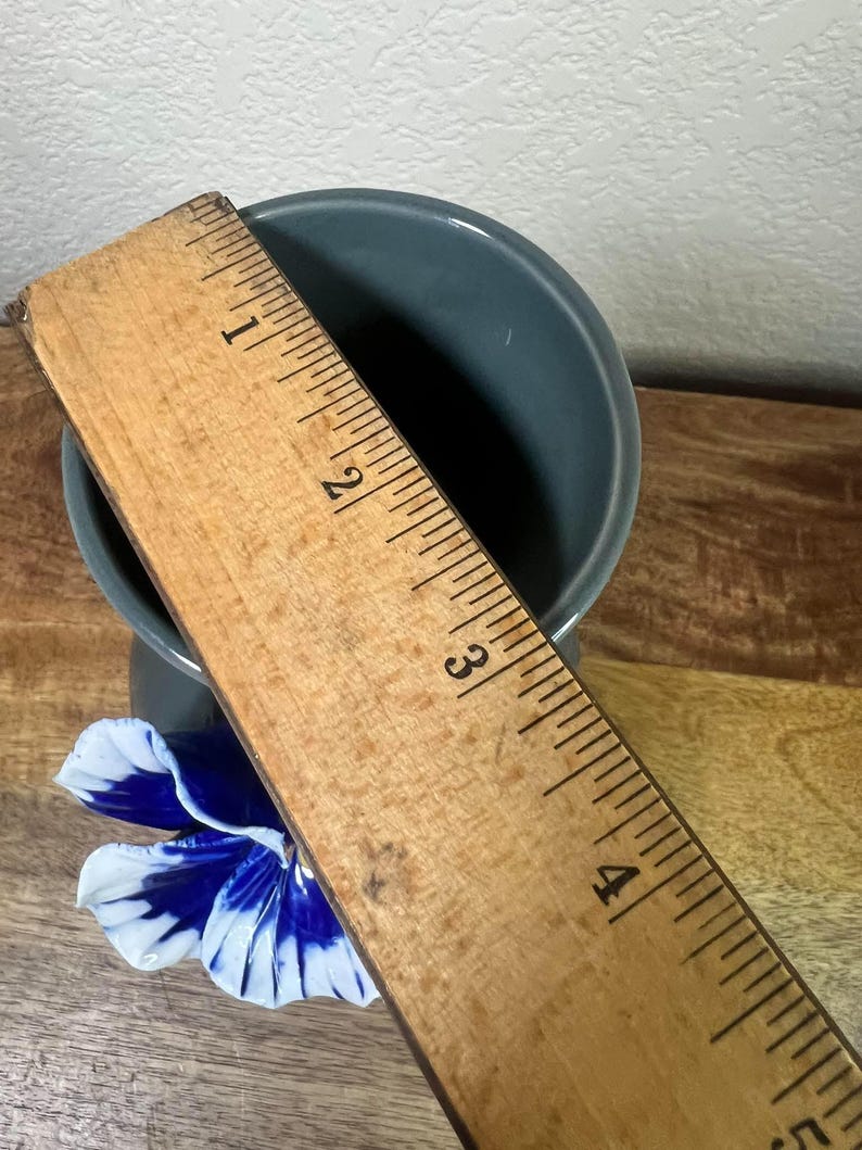 May include: A small, teal-colored ceramic vase with a blue and white flower accent. A wooden ruler is placed over the vase, measuring approximately 4 inches in height. The vase sits on a wooden surface.