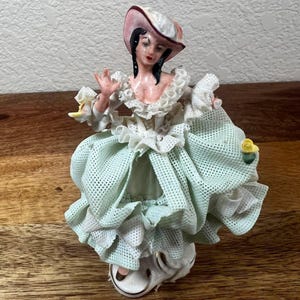 May include: A porcelain figurine of a person wearing a wide-brimmed hat and a voluminous, light green dress with a textured, grid-like pattern. The dress features white ruffled lace trim at the neckline and sleeves, accented with small yellow flowers.