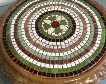 May include: A round, wooden table top with a mosaic design. The mosaic is made of small, square tiles in shades of green, brown, and white. The center of the mosaic features a stylized flower with white petals and a red center.
