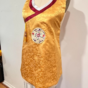 May include: A gold sleeveless dress with a floral pattern and a red and white trim. The dress has a round embroidered patch on the front with a floral design. The dress is on a mannequin.