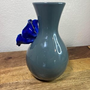 May include: A gray ceramic vase with a vibrant blue flower accent. The vase has a rounded base and a flared top. The flower is attached to the side of the vase, adding a decorative touch. The vase is sitting on a wooden surface.