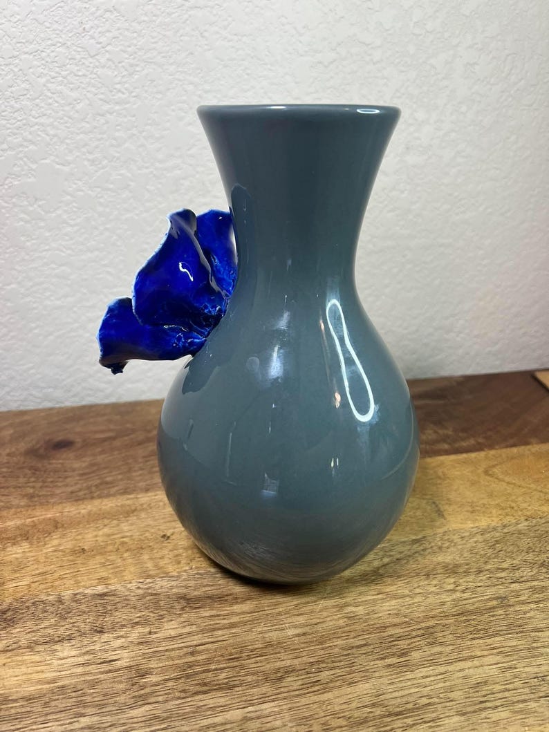 May include: A gray ceramic vase with a vibrant blue flower accent. The vase has a rounded base and a flared top. The flower is attached to the side of the vase, adding a decorative touch. The vase is sitting on a wooden surface.