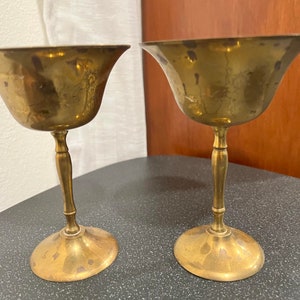 Puede incluir: Dos copas de metal de color dorado con un cuenco ancho y un tallo alto y delgado. Las copas están sobre una superficie oscura.