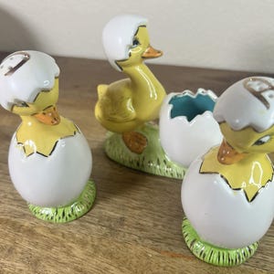 May include: Set of four ceramic salt and pepper shakers shaped like ducklings in eggshells. The ducklings are yellow with black eyes and beaks. The eggshells are white with gold accents.