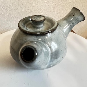 May include: A handmade ceramic teapot with a round body and a curved spout. The teapot is light gray with darker gray speckles and a dark brown rim. It has a lid with a small knob. A unique piece for tea lovers.