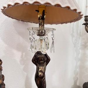 May include: A table lamp with a pink lampshade and a crystal base. The base features a cherub sculpture holding a crystal bowl with hanging crystal drops.