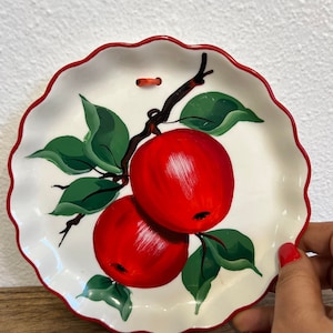 Peut inclure: Une assiette en céramique blanche avec un bord rouge et une bordure festonnée. L'assiette présente un motif peint à la main de deux pommes rouges sur une branche avec des feuilles vertes.