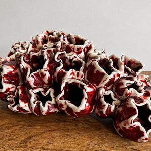 Pode incluir: Uma escultura em cerâmica com um conjunto de formas orgânicas texturizadas. A peça apresenta um esquema de cores vermelho escuro e branco, com o vermelho aparecendo em um padrão marmorizado. A escultura é exibida em uma superfície de madeira.