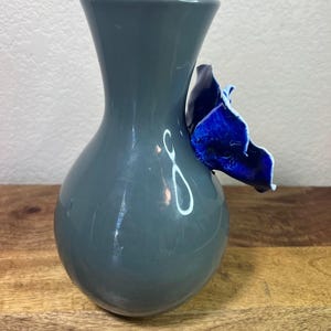 May include: A gray ceramic vase with a bulbous body and a flared neck. A vibrant blue, petal-shaped accent is attached to the side. The vase is displayed on a wooden surface.