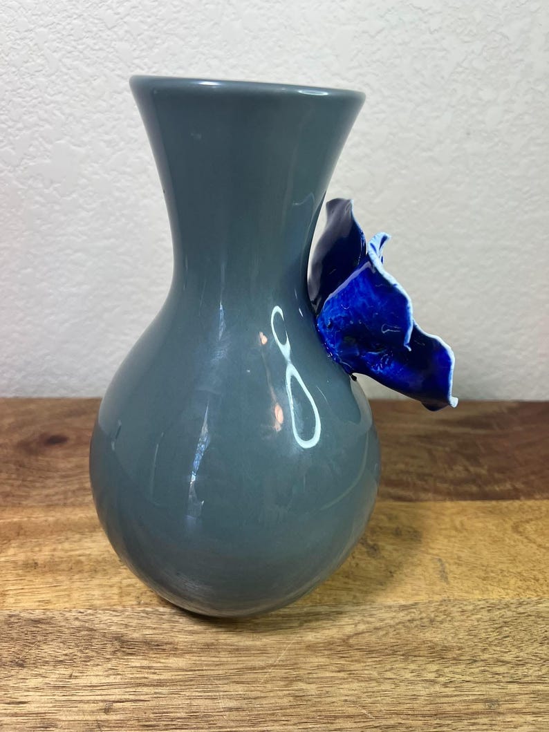 May include: A gray ceramic vase with a bulbous body and a flared neck. A vibrant blue, petal-shaped accent is attached to the side. The vase is displayed on a wooden surface.