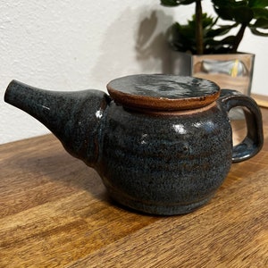 Op de afbeelding: Een klein, donkerblauw aardewerken theepot met een bruin deksel. De theepot heeft een tuit en een handvat. De theepot staat op een houten oppervlak.