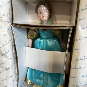 May include: A porcelain doll wearing a blue and gold dress. The doll is in a box with the words "The Danbury Mint" printed on the sides.