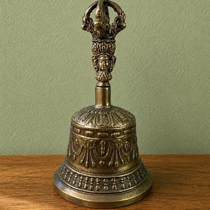 May include: A detailed bronze bell with intricate carvings. The bell features a decorative handle with a detailed face and crown design. The bell's body has ornate patterns and a rounded base, showcasing a vintage aesthetic.