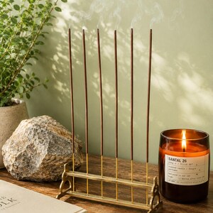 May include: A gold-colored incense holder with six burning incense sticks, emitting wisps of smoke. A lit candle in a glass jar is to the right, and a book is in the foreground. The candle label reads "SANTAL 26".