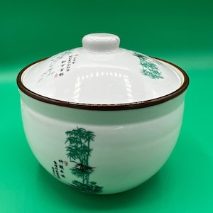 Könnte beinhalten: Weiße Keramikschüssel mit Deckel, mit braunem Rand und einem grünen Bambusdesign mit chinesischen Schriftzeichen. Die Schüssel steht auf einem grünen Hintergrund.