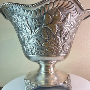 May include: A silver bowl with a floral design and a scalloped rim. The bowl sits on a pedestal with three feet.