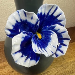 May include: A ceramic vase with a large, decorative flower. The flower has white petals with blue accents and a yellow center. The vase is dark gray and sits on a wooden surface.