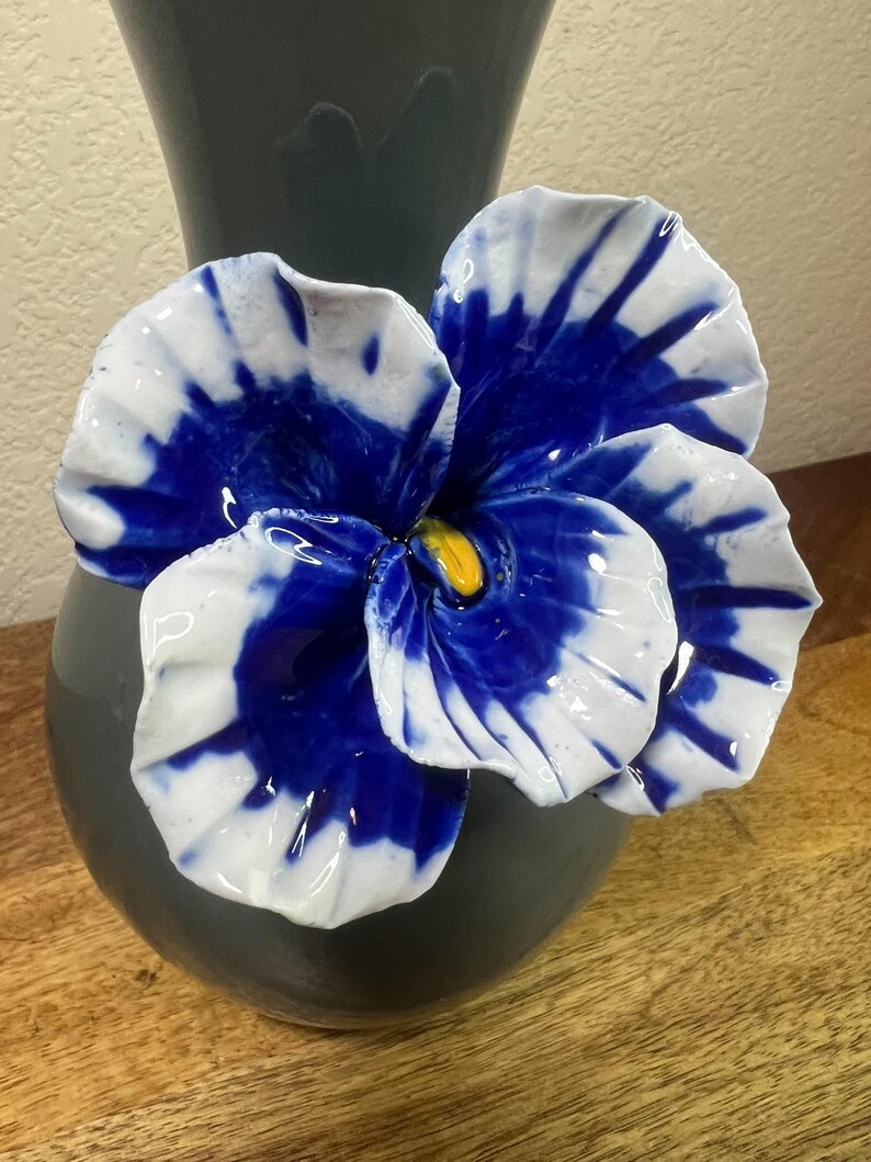 May include: A ceramic vase with a large, decorative flower. The flower has white petals with blue accents and a yellow center. The vase is dark gray and sits on a wooden surface.