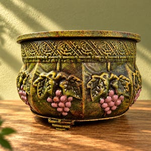 May include: A round, footed planter featuring a textured, moss-green finish with embossed grape clusters in a soft pink hue. The upper rim displays a decorative geometric pattern, while the body is divided into panels showcasing sculpted grape leaves.