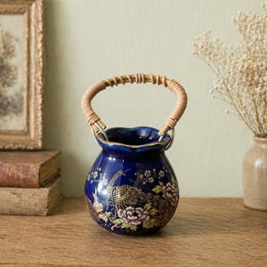 May include: A dark blue ceramic vase with a woven handle. The vase features a floral and peacock design in gold, pink, and green. The rim is trimmed with gold. The vase is on a wooden surface.