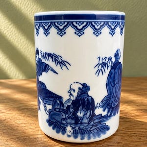 May include: A white ceramic mug featuring a blue hand-painted scene of figures in traditional attire sitting near bamboo stalks. The top rim is decorated with a repeating blue scalloped pattern. The mug has a cylindrical shape and a smooth finish.