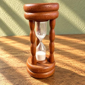 May include: An hourglass featuring a warm-toned brown wooden frame with a circular top and base connected by four turned spindles. A clear glass bulb is positioned in the center, containing fine white sand that is currently flowing downward.