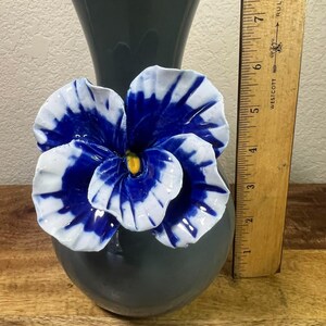 May include: A gray ceramic vase with a large, three-dimensional blue and white flower attached. The flower has a yellow center. A wooden ruler is placed next to the vase, showing measurements in inches.