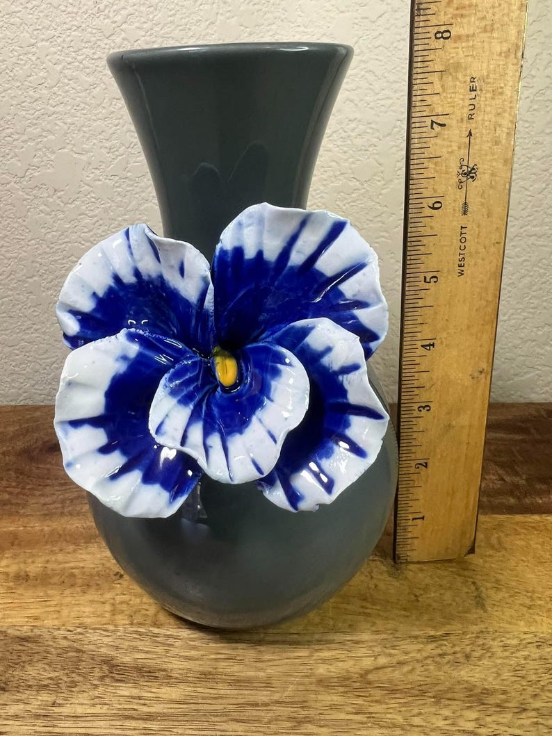 May include: A gray ceramic vase with a large, three-dimensional blue and white flower attached. The flower has a yellow center. A wooden ruler is placed next to the vase, showing measurements in inches.