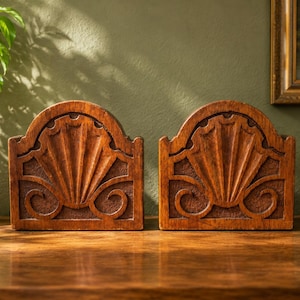 May include: A pair of matching wooden bookends featuring a carved shell motif. Each piece has a semi-circular top edge, a central fan-shaped scallop shell design, and symmetrical scrollwork at the base, all finished in a medium brown wood tone.