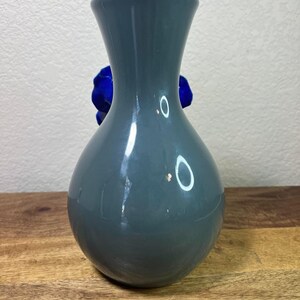 May include: A gray-blue ceramic vase with a rounded body and flared neck. A vibrant blue flower is visible behind the vase. The vase is displayed on a wooden surface.